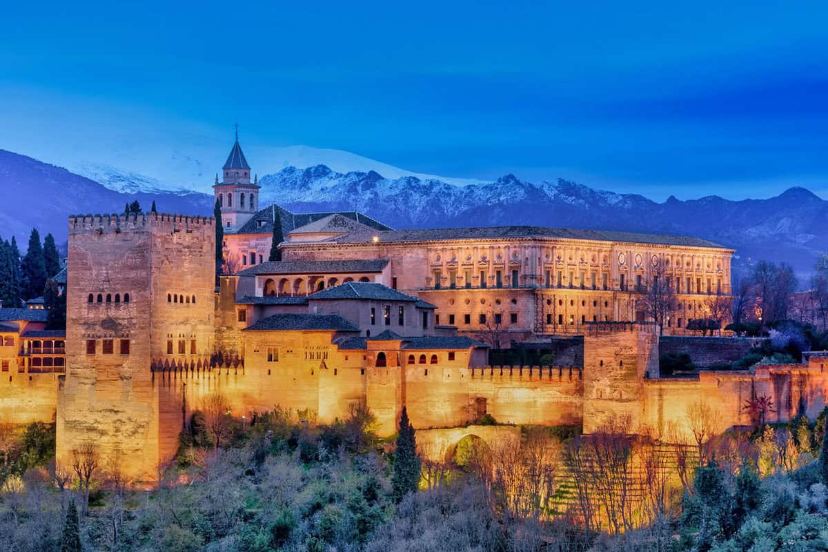 Scenic view of the Alhambra palace in Granada, Spain, illuminated at dusk with the Sierra Nevada mountains in the background.