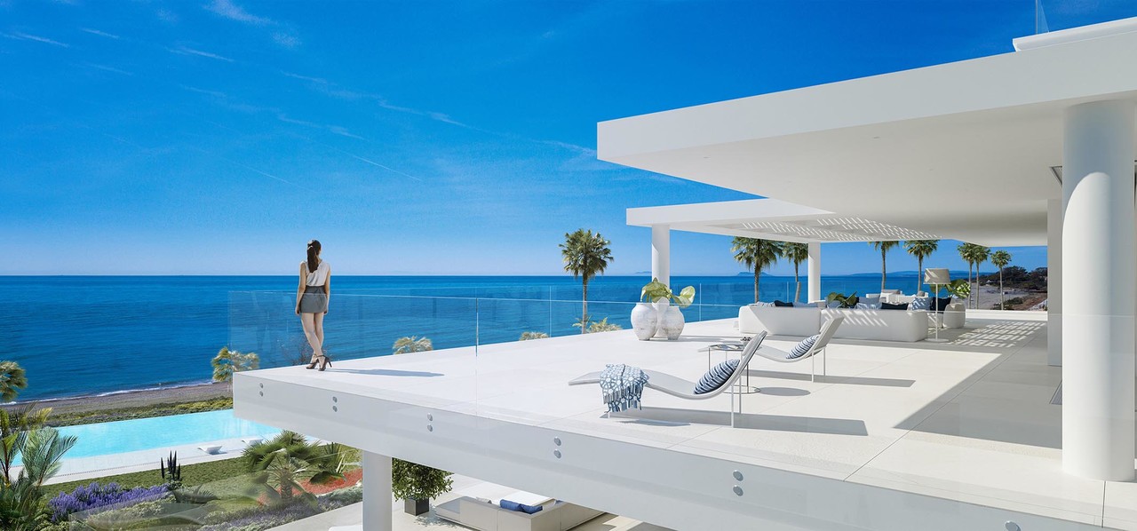 Luxury oceanfront terrace with modern white architecture, lounge chairs, and a woman enjoying the panoramic sea view under a bright blue sky.